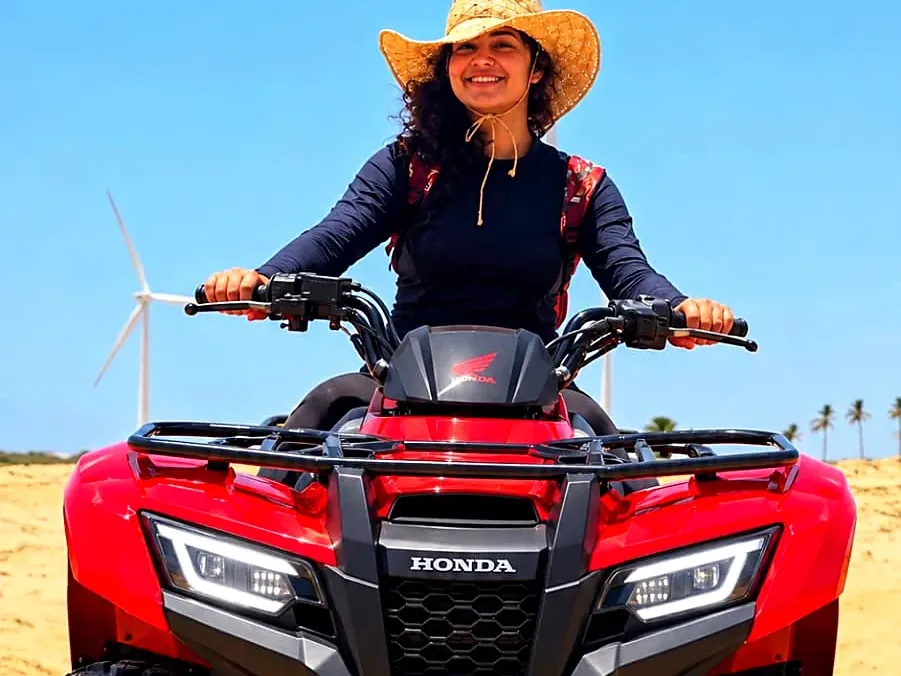Visitante sorrindo sobre um quadriciclo vermelho na areia dos Pequenos Lençóis, com os cataventos ao fundo